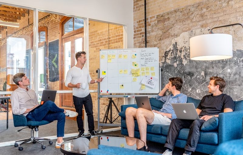 three men sitting while using laptops and watching man beside whiteboard